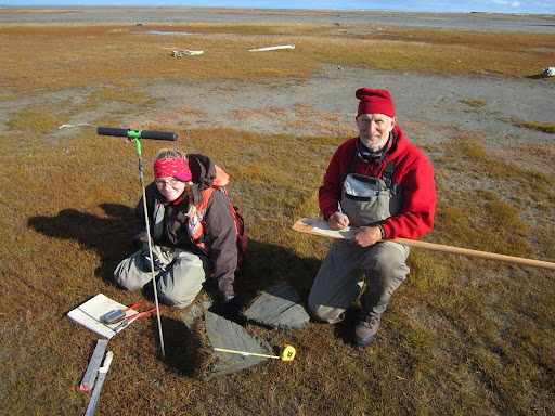 Two heavily clothed people kneel on wet tundra with instruments and notepads.